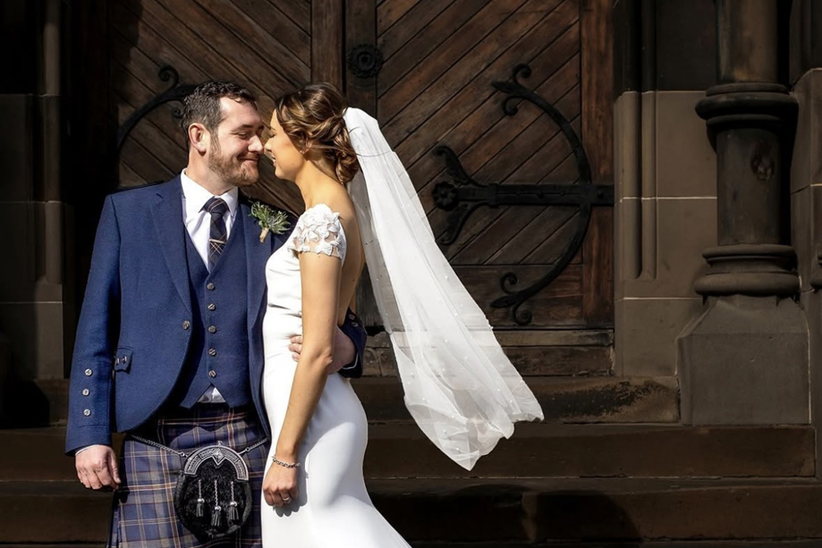 Bride wearing veil and groom wearing a kilt standing outside venue in an embrace while smiling at each other